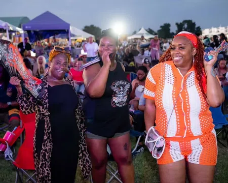Three smiling women at an outdoor event with tents and crowd in the background during evening.