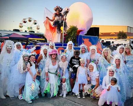 Group of people in white costumes and wings posing outdoors at a festival with a ferris wheel and colorful float.