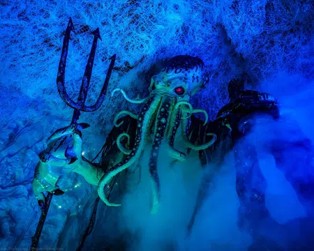 Cthulhu figure with glowing red eyes holding a trident amidst blue fog and web-like textures underwater.