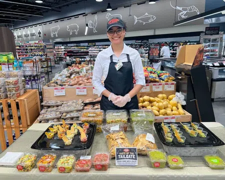 Smiling store employee presenting various homemade dips and salsas at a grocery store sampling table.