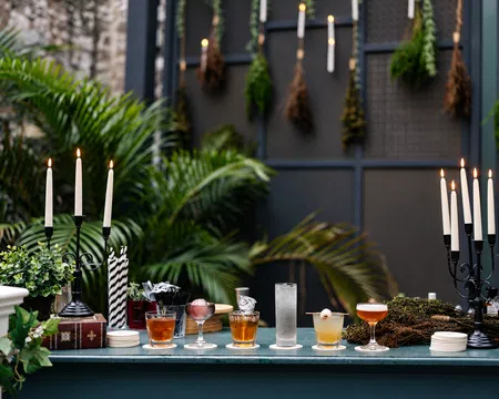 Outdoor bar setup with assorted cocktails, lit candles, greenery, and dried herbs hanging on a dark backdrop.