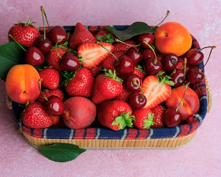 Basket filled with fresh strawberries, cherries, peaches, and apricots on a pink surface