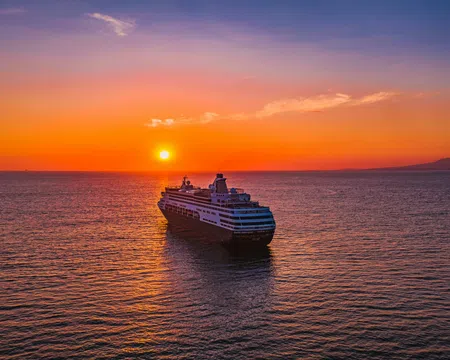 Large cruise ship sailing on calm ocean waters during vibrant orange sunset with clear sky.