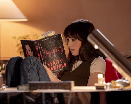 Woman reading the book Hospitality by Verity Crawford in a cozy, warmly lit room with a table lamp.