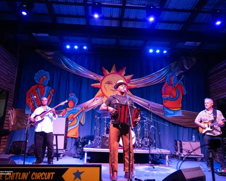 Live band performing on stage with accordion, guitar, and bass under bright lights and colorful sun backdrop