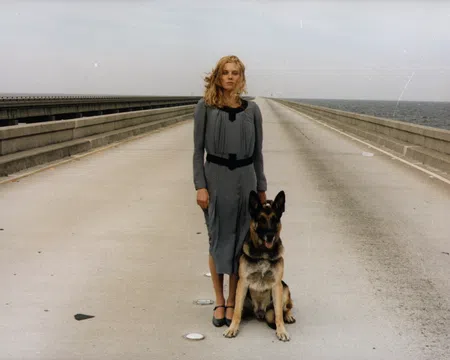 Woman in gray dress stands with German Shepherd on empty bridge over water under cloudy sky.