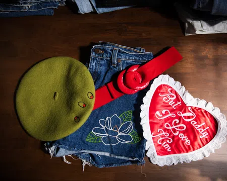 Green beret, denim shorts with floral embroidery, red belt, and heart-shaped pillow with love message on wooden surface.