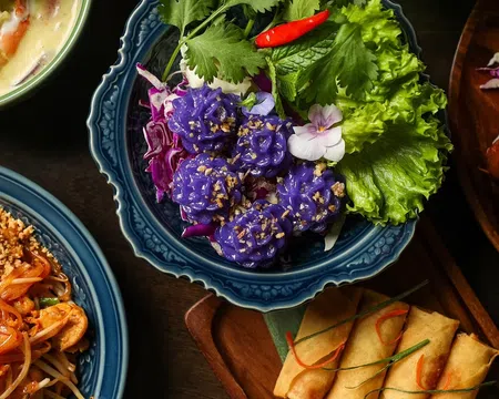 Colorful Thai dishes including purple flower-shaped dumplings, spring rolls, pad thai, and curry on ornate plates.