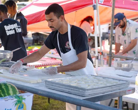 Man wearing apron preparing dough at outdoor food stall with trays of dough and tents in background