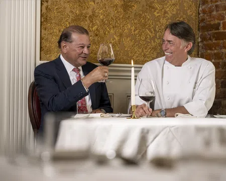 Two men smiling and toasting with red wine in an elegant dining room with gold wallpaper and exposed brick.