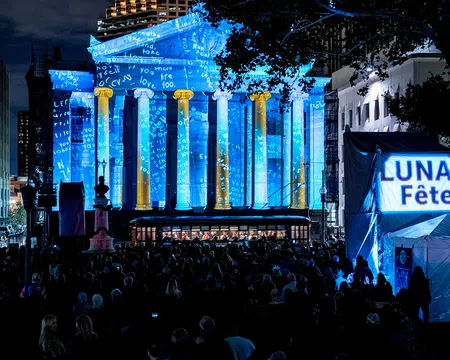 Crowd at night watching blue and gold light projections on a classical building during Luna Fête festival.
