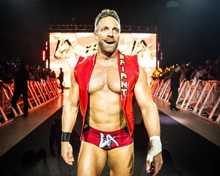 Muscular male wrestler in red trunks and vest walking confidently toward ring with audience and lights behind him
