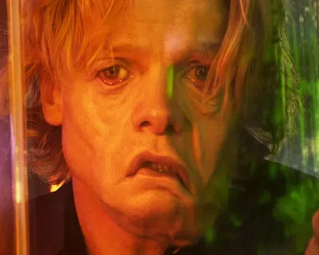 Close-up of a distressed man with blonde hair looking through a glass panel illuminated by orange and green light.