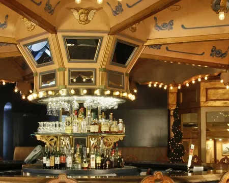 Elegant vintage bar interior with ornate ceiling, lights, central liquor display, and wooden chairs under warm lighting.