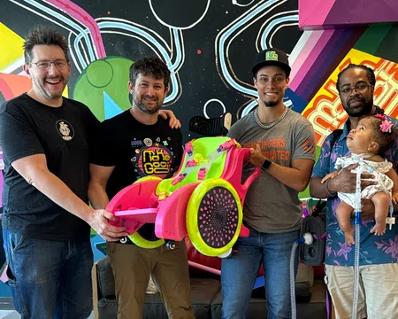 Group of five people smiling indoors with colorful wall art, holding a bright pink and green custom wheelchair.