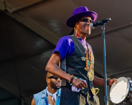 Musician in purple hat and vest singing into microphone while playing tambourine on stage at outdoor event.