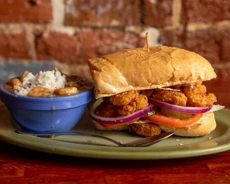 Crispy fried shrimp po' boy sandwich with lettuce, tomato, onion, served with a side of rice and beans in a blue bowl.