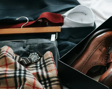 Flat lay of men's brown dress shoes in a box, plaid scarf, gray trousers, watch, and a white shirt on a dark suit jacket.