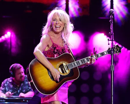 Female country singer in pink dress playing acoustic guitar on stage with vibrant purple lighting and smiling joyfully.