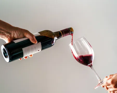 Red wine being poured from a bottle into a glass held by two hands against a plain background.