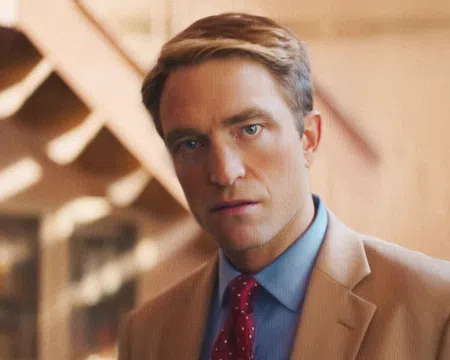 Man in a tan suit and red polka dot tie staring directly at the camera with a blurred warm background.