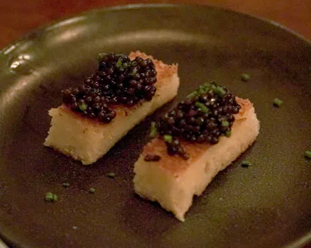 Two rectangular pieces of toasted bread topped with black caviar and garnished with chopped chives on a dark plate