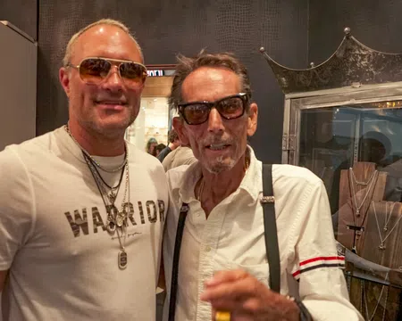 Two men wearing sunglasses and necklaces posing indoors at a jewelry store with display cases behind them
