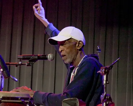 Elderly male musician playing congas and singing into a microphone with one hand raised on stage.