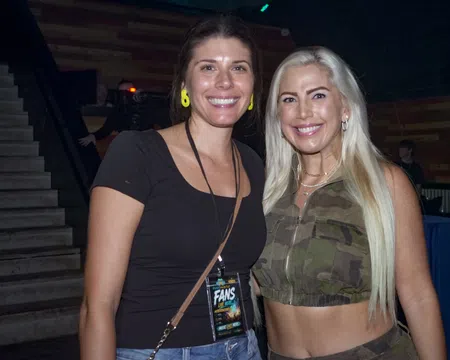 Two smiling women posing indoors, one in black top and the other in camouflage crop top with long blonde hair.