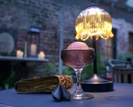 Elegant cocktail glass with a pink spherical garnish on a table beside a decorative lamp and fortune cookies.