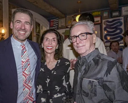Three adults smiling together indoors, dressed in semi-formal attire at a social gathering with framed art on walls.