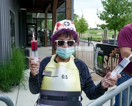 Person in nurse costume wearing sunglasses and mask holding two large syringes outside a building.