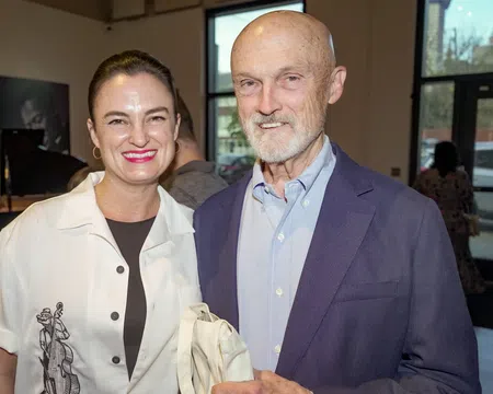 Smiling woman and elderly man standing indoors at a social event with large windows in the background.