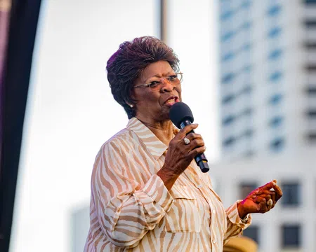 Elderly woman in striped blouse speaking into microphone at outdoor event with city building background