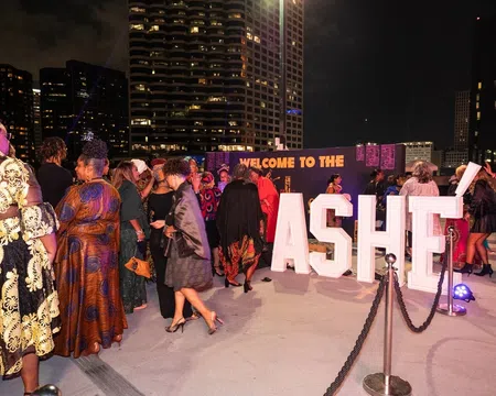 People dressed in elegant attire gather at a nighttime outdoor event with large illuminated ASHE letters and city buildings in the background