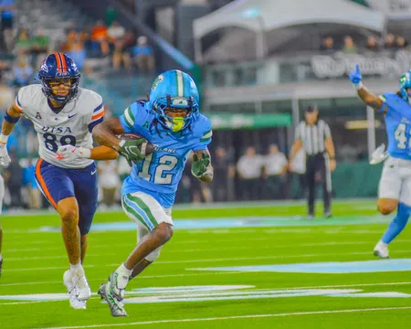 Football player in blue uniform running with ball as opponent in white uniform chases him on the field.