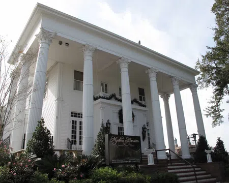 White Greek Revival mansion with tall Corinthian columns and manicured garden in front under a clear sky.
