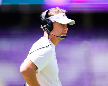 Football coach wearing LSU visor and headset focused during a game with blurred purple stadium background