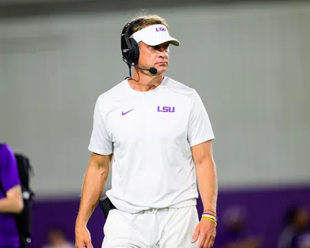 LSU football coach in white LSU gear and visor wears a headset on the sidelines during a game.