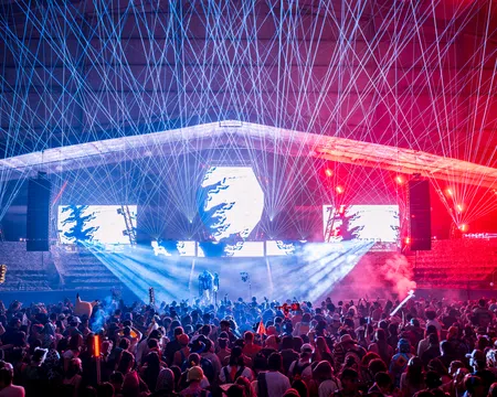 Crowd enjoying a nighttime concert with vibrant laser lights and colorful stage effects under a large canopy.