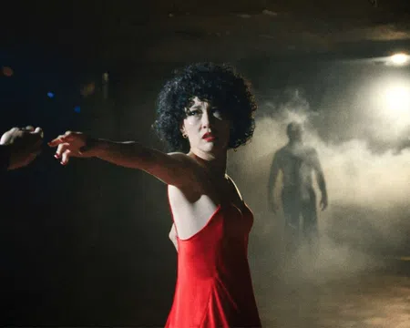 Woman in a red dress reaching out hand in a dark room with mist and a shadowy figure in the background