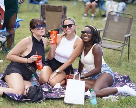 Three women wearing sunglasses enjoying drinks while sitting on a blanket at an outdoor event on grass.