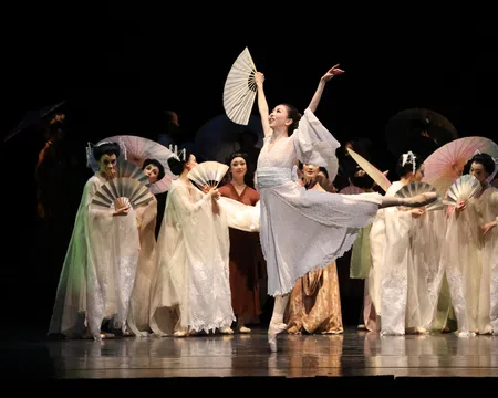 Ballet dancer in white costume performs with fans on stage with other dancers in traditional attire.