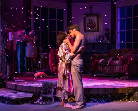 Couple embraces on stage in intimate theater scene with warm lighting and scattered petals.