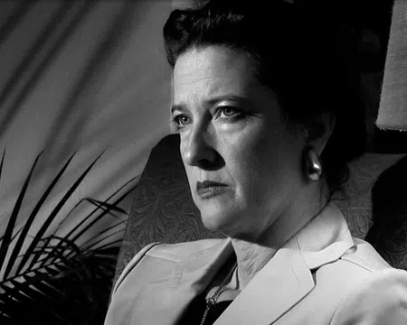 Black and white portrait of a serious woman sitting in a chair with shadows and a lamp in the background