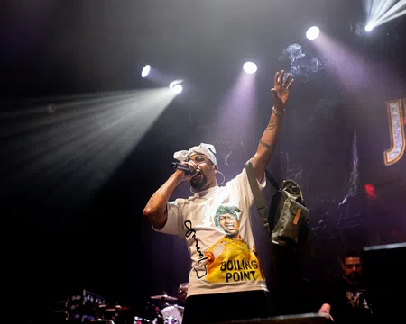 Hip hop artist performing on stage under bright spotlights with microphone and raised arm during concert.