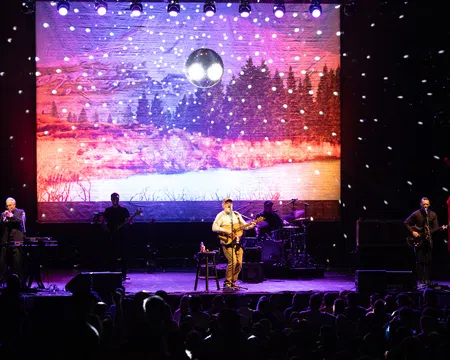 Band performing on stage with snow effects and forest landscape backdrop under disco ball lighting.