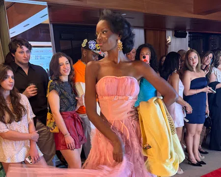 Model in pink gown holding yellow jacket walking past crowd of spectators at indoor event with wood paneling.