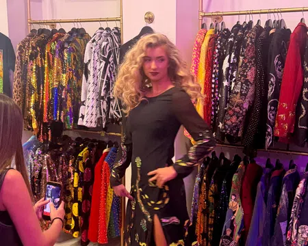 Woman in black dress posing in a colorful boutique with sequined and patterned jackets on racks
