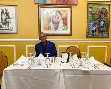 Man in blue suit sitting at white tablecloth-covered table set with wine glasses and silverware, framed art on yellow wall.